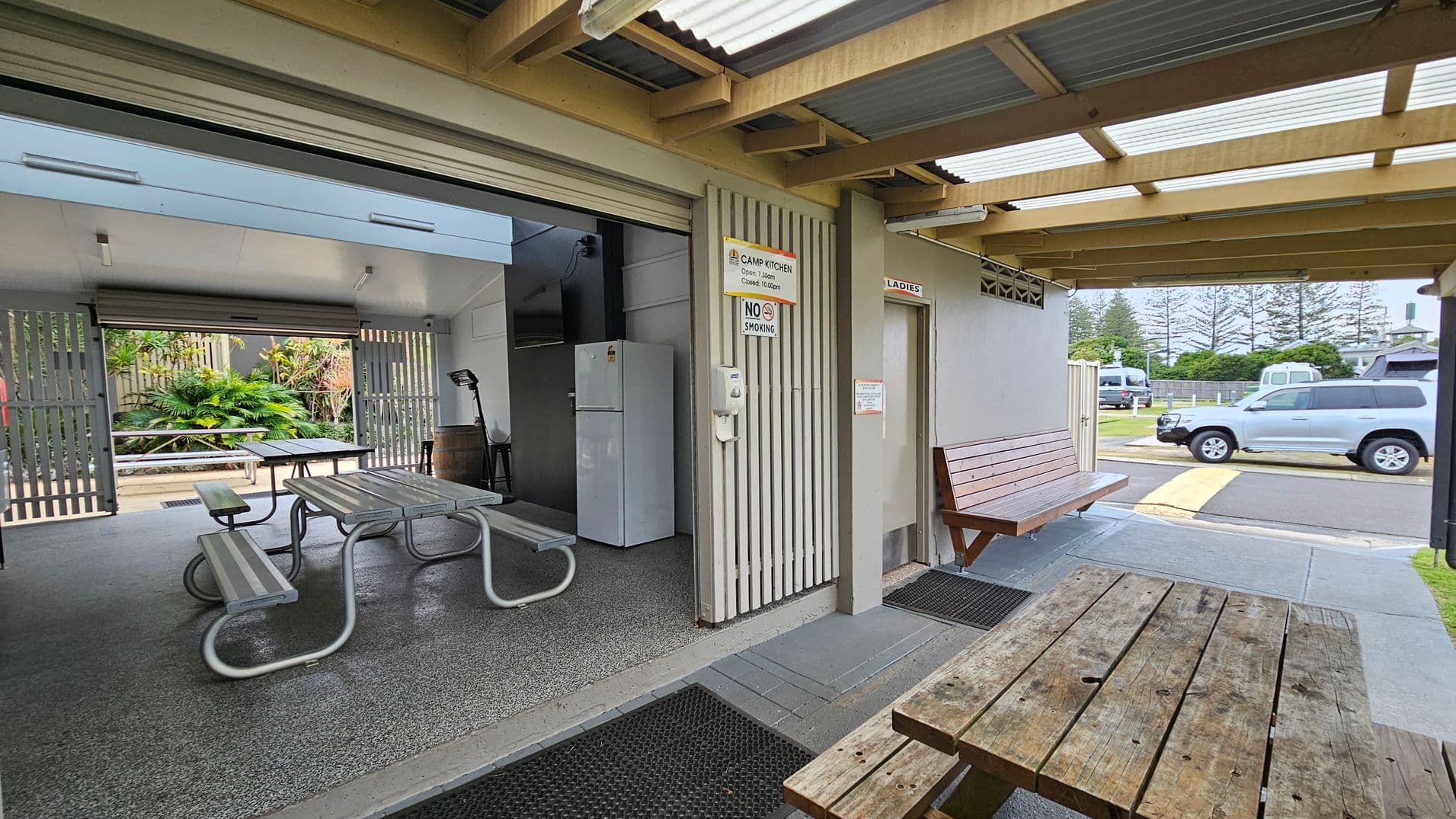 A covered outdoor area with a wooden picnic table in the foreground. To the left, there is an open camp kitchen with metal picnic tables and a white refrigerator. The kitchen has a grey, speckled floor and a partially open roller door. A "No Smoking" sign is visible. To the right, a wooden bench is positioned outside a door marked "Ladies." The ceiling is made of corrugated metal with wooden beams. In the background, a road with parked vehicles and trees is visible.