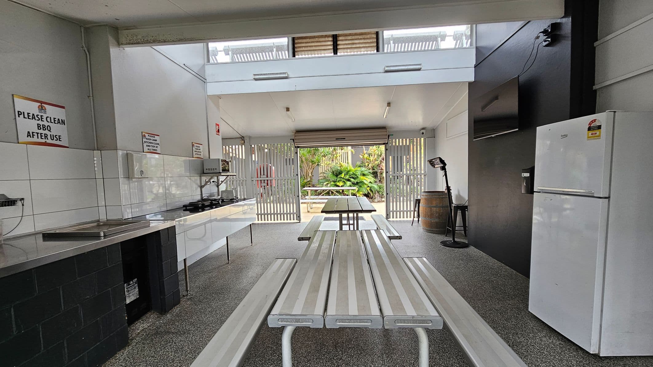 A covered outdoor kitchen area with a long picnic table and benches in the centre. On the left, there is a countertop with a sink and a gas stove, accompanied by signs reminding users to clean the BBQ. On the right, a white refrigerator stands against a dark wall, with a TV mounted above. The floor is speckled grey, and the ceiling is white with lighting fixtures. The background features a garden with lush greenery visible through a slatted fence.