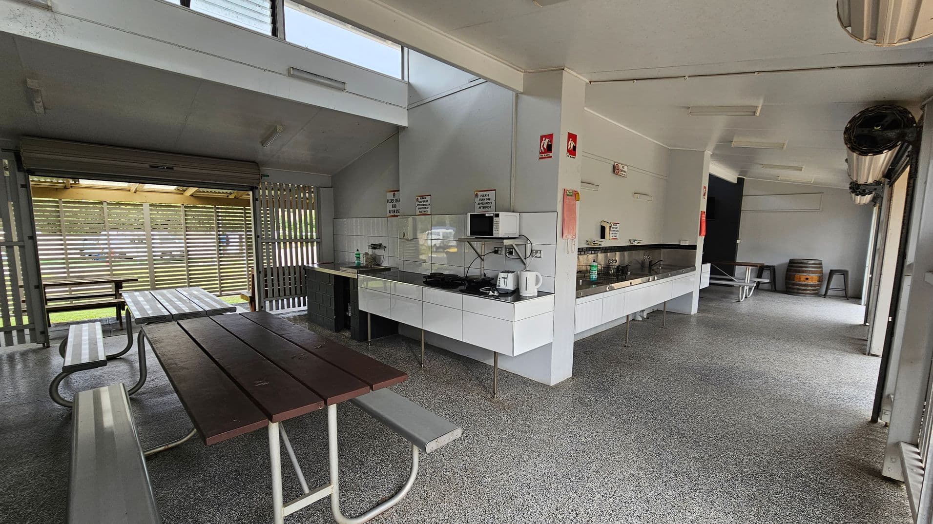 An indoor picnic area with a speckled grey floor and a white ceiling. On the left, there are picnic tables with attached benches. The background features a slatted wooden wall with a view of grass outside. On the right, a kitchen area with white cabinets, a microwave, a kettle, and a sink. A barrel and stools are visible further back. The space is well-lit with fluorescent lights and has a modern, functional design.