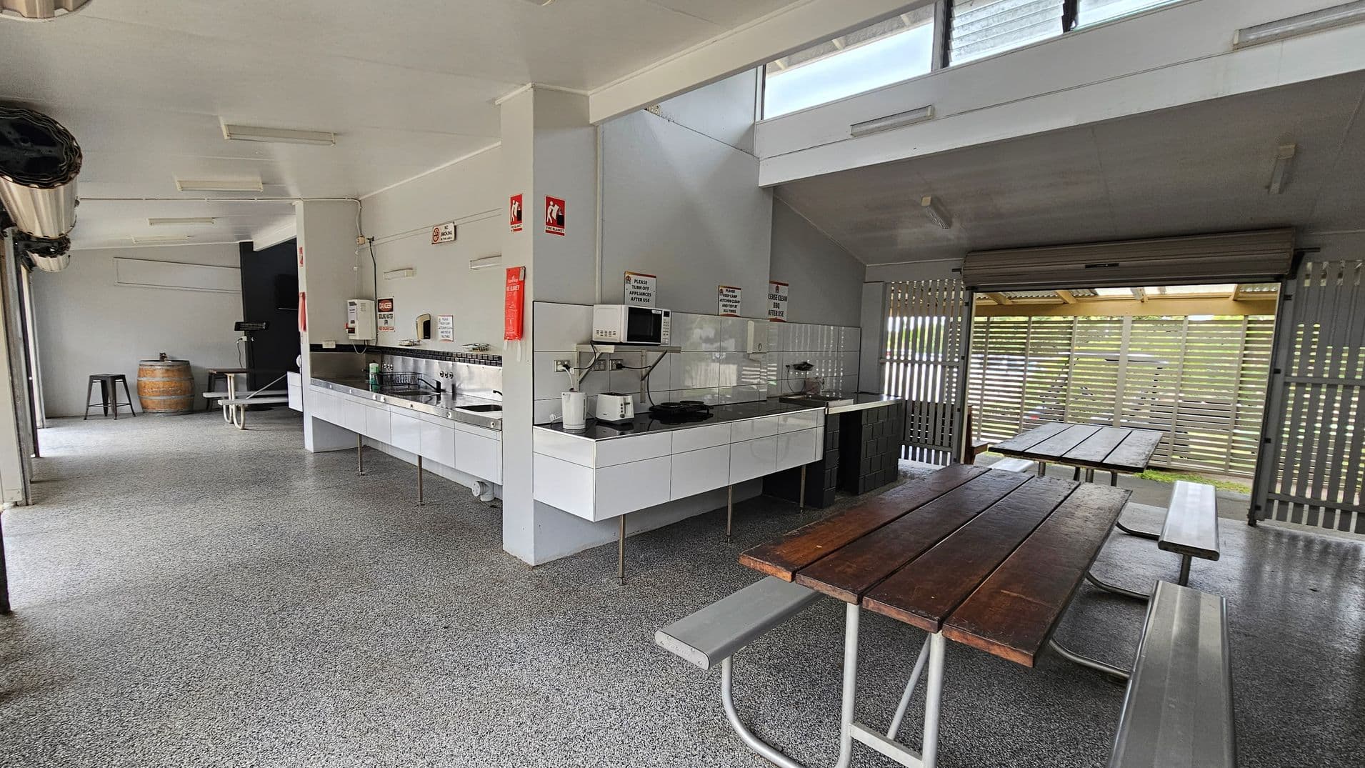 A spacious indoor kitchen area with a speckled grey floor and a light-coloured ceiling. On the right, there are wooden picnic tables with metal benches. In the centre, a long counter with white cabinets, a microwave, and various kitchen appliances. On the left, a barrel and stools are visible. The back wall has a large opening with slatted wooden panels, allowing natural light to enter. Signs and safety equipment are mounted on the walls.