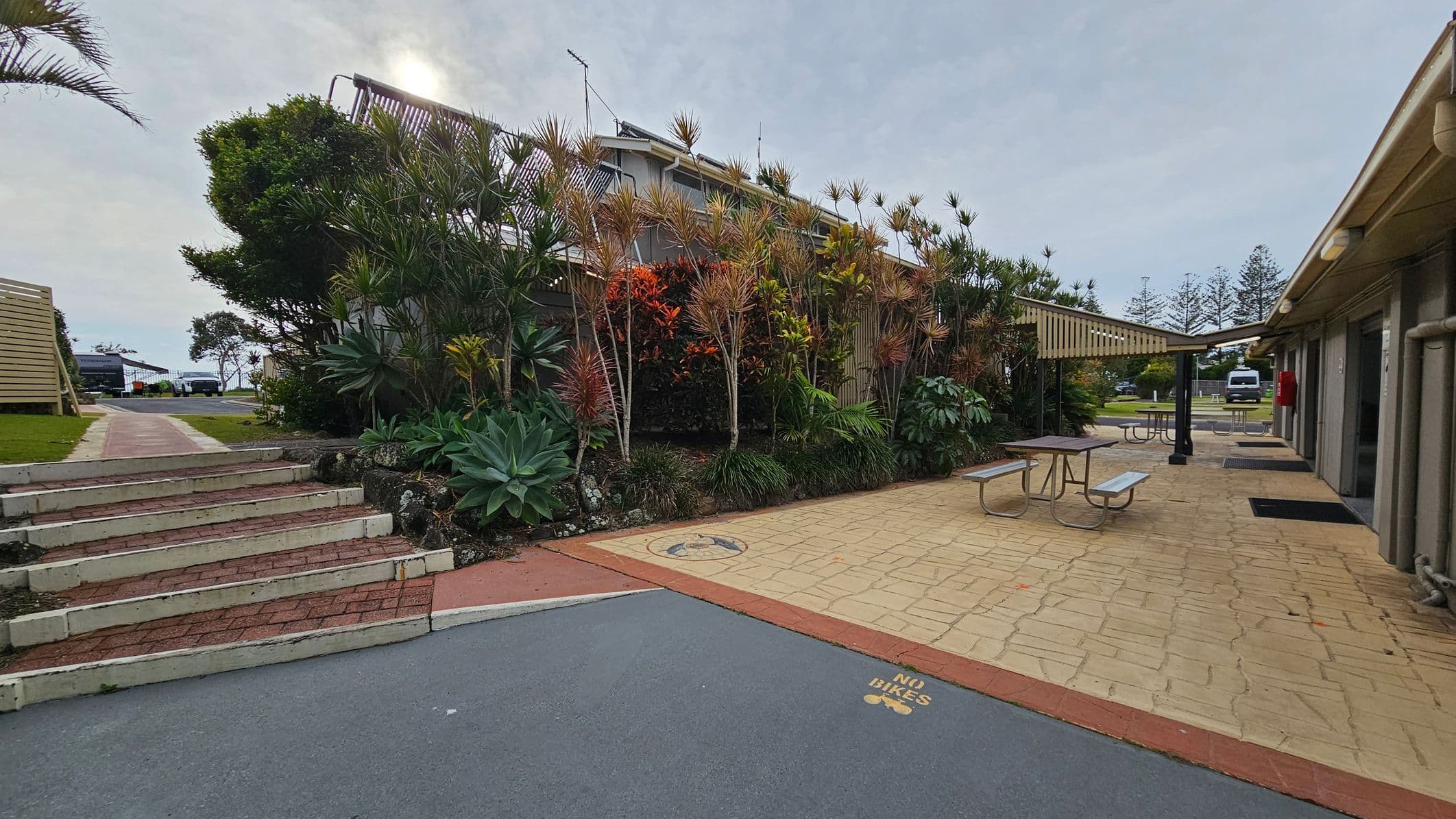 On the left, there are steps leading up to a path bordered by a wooden fence and lush greenery, including large succulents and tall plants. In the centre, a garden with various plants and a small tree is visible. On the right, a paved area with picnic tables under a pergola is adjacent to a building with a sloped roof. A "No Bikes" sign is painted on the ground. In the background, there are more trees and a parked vehicle.