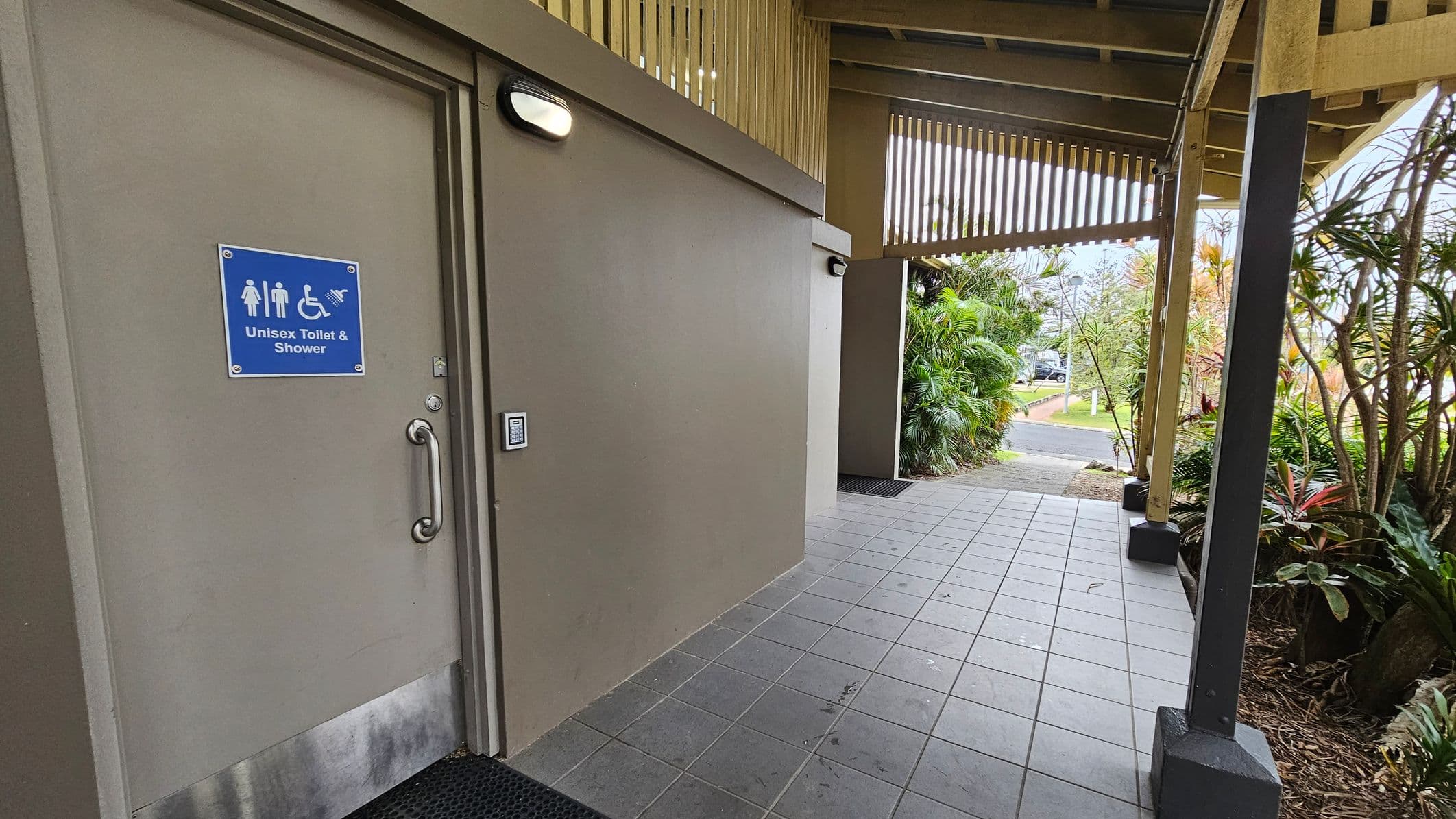 A tiled walkway with a unisex toilet and shower door on the left, featuring a blue sign with symbols for male, female, and disabled access. The door has a silver handle and a keypad entry. On the right, there are lush green plants and a wooden structure with vertical slats. The ceiling is wooden with exposed beams. The path leads to an outdoor area with more greenery and a glimpse of a parked car in the distance.