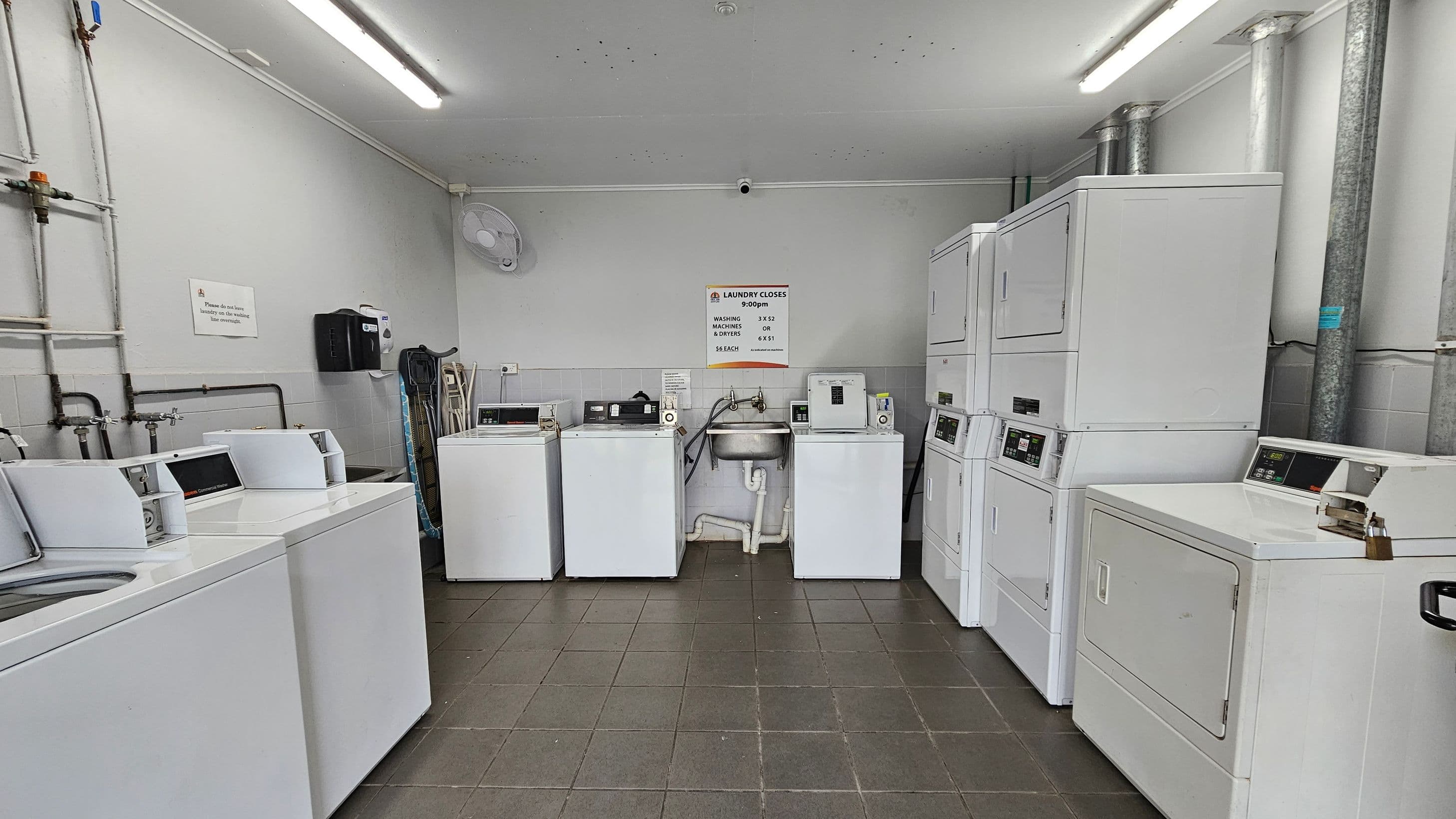A laundry room with several white washing machines and dryers. On the left, there are three top-loading washing machines. In the centre, a stainless steel sink is flanked by two more machines. On the right, there are stacked dryers and another machine. The walls are light grey with visible pipes and a wall-mounted fan. A sign with laundry instructions is above the sink. The floor is tiled in dark grey, and the ceiling has fluorescent lights.