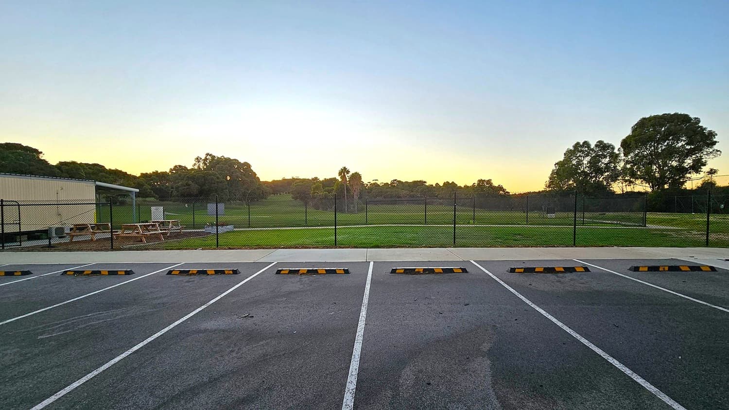 A row of empty parking spaces with yellow and black wheel stops in the foreground. Beyond the parking area, a chain-link fence encloses a grassy field. To the left, there are picnic tables and a small structure. Trees are scattered across the background, with a few taller ones silhouetted against the sky. The ground is paved, and the sky has a gradient from light to darker blue.