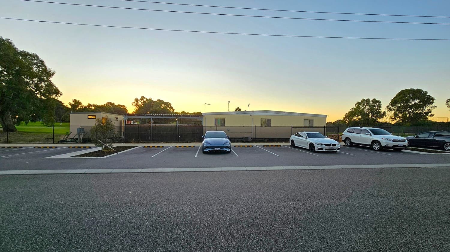 A parking area with several cars lined up. In the centre, a blue car is parked, flanked by a white car on the right and another white vehicle further to the right. On the far right, a dark-coloured car is visible. In the background, a single-storey building with a flat roof and small windows is surrounded by a fence. Trees are visible on both sides, with a grassy area on the left. The ground is paved, and there are parking space markers.