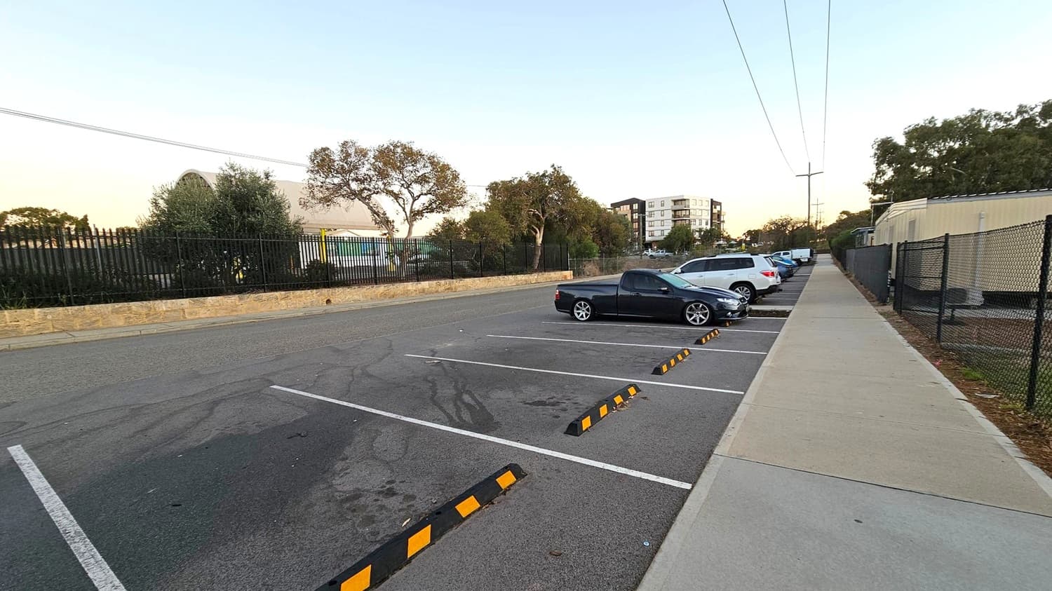 A parking area with several cars parked on the right side, including a black vehicle in the foreground. The left side features a stone wall and a black metal fence with trees and shrubs behind it. In the background, a multi-storey building is visible. A footpath runs parallel to the parking spaces on the right, bordered by a black chain-link fence. Overhead, power lines stretch across the scene. The ground is asphalt, and the footpath is concrete with a green strip.