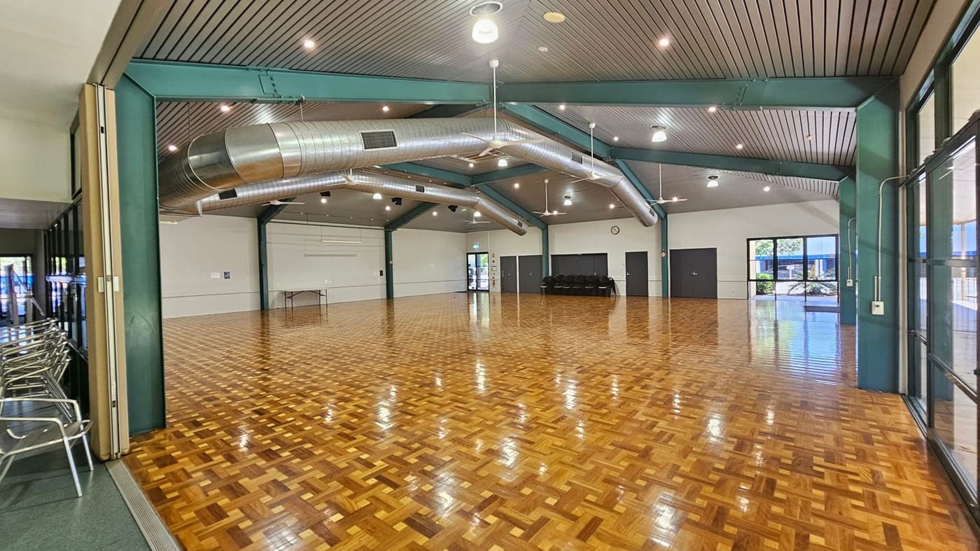 A spacious hall with a polished parquet floor and a high ceiling featuring exposed metal beams and large air ducts. On the left, there are stacked metal chairs against a wall. The back wall has a clock and several doors, with a small table nearby. Large windows on the right allow natural light to enter, showing a glimpse of greenery outside. The ceiling has multiple lights and fans.
