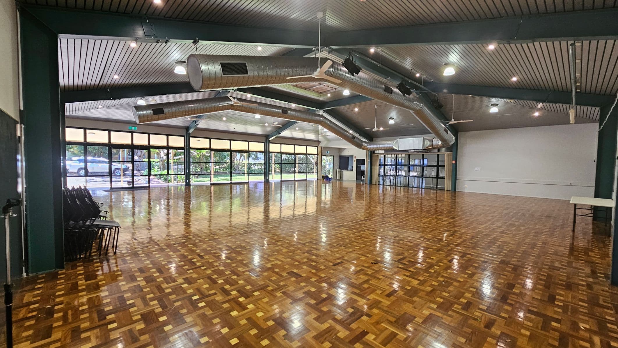 A spacious hall with a polished parquet floor. On the left, there are stacked chairs against the wall. The right side features a plain white wall with a small table. The background has large windows and glass doors, allowing natural light to enter and providing a view of greenery and parked cars outside. The ceiling is lined with exposed ductwork and lighting fixtures.