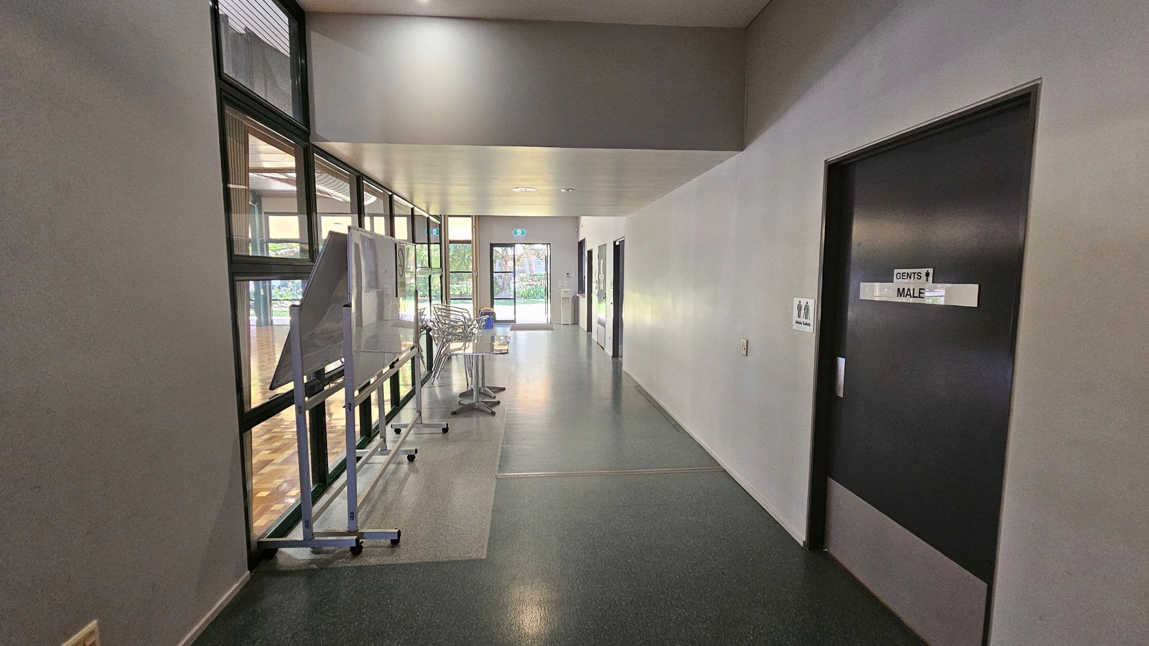 A hallway with a dark floor and light ceiling. On the right, there's a door marked "Gents Male" with a sign next to it. On the left, large windows reveal a wooden-floored room. A whiteboard and stacked chairs are positioned along the windows. At the end of the hallway, a glass door leads outside, with greenery visible through it.