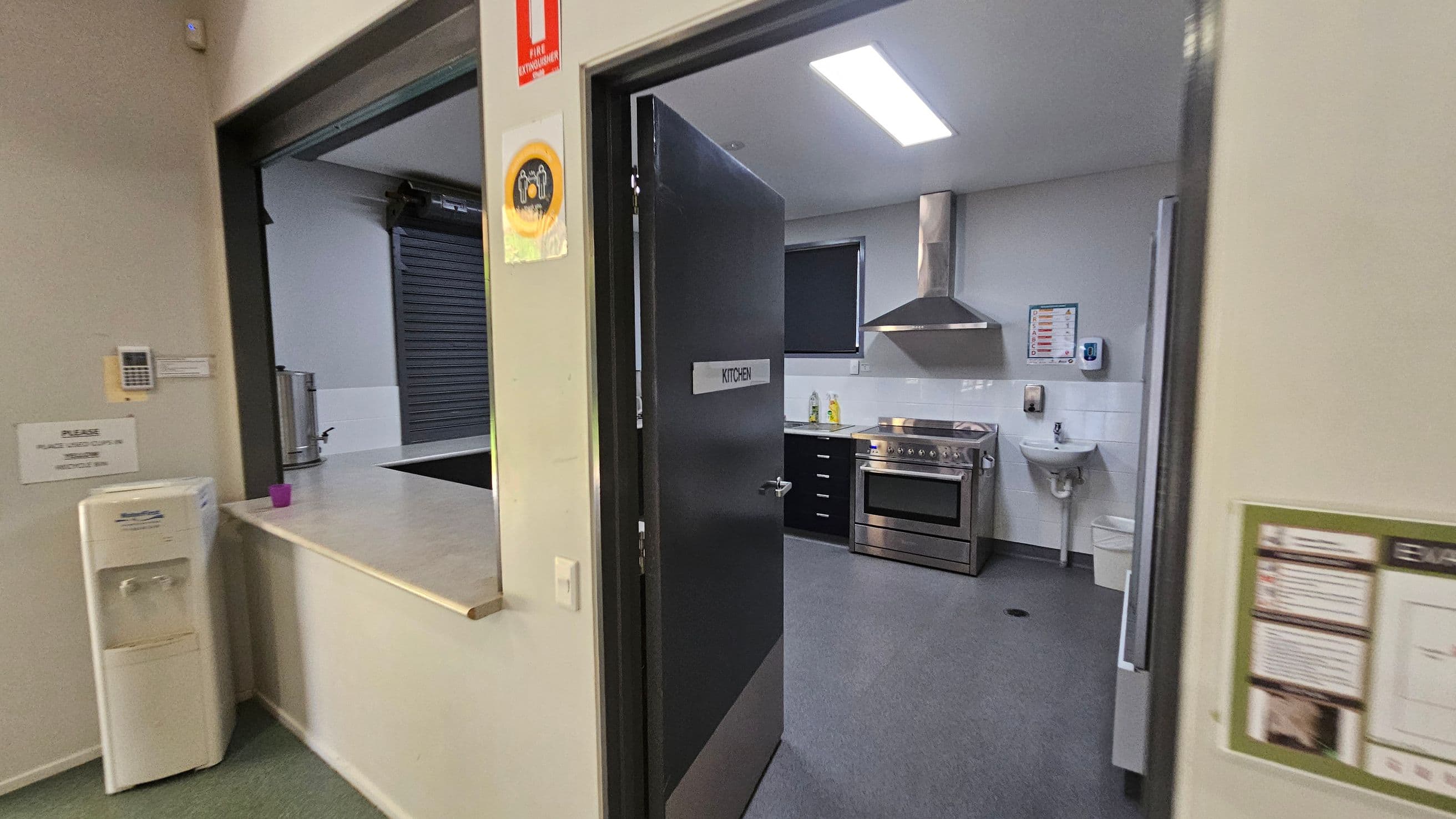 A kitchen with a black door labelled "KITCHEN" is open, revealing a stainless steel oven and range hood. A small sink is mounted on the wall to the right. On the left, a counter with a water cooler and a purple cup is visible. A fire extinguisher sign is above the door. The floor is grey, and the ceiling has a bright fluorescent light.
