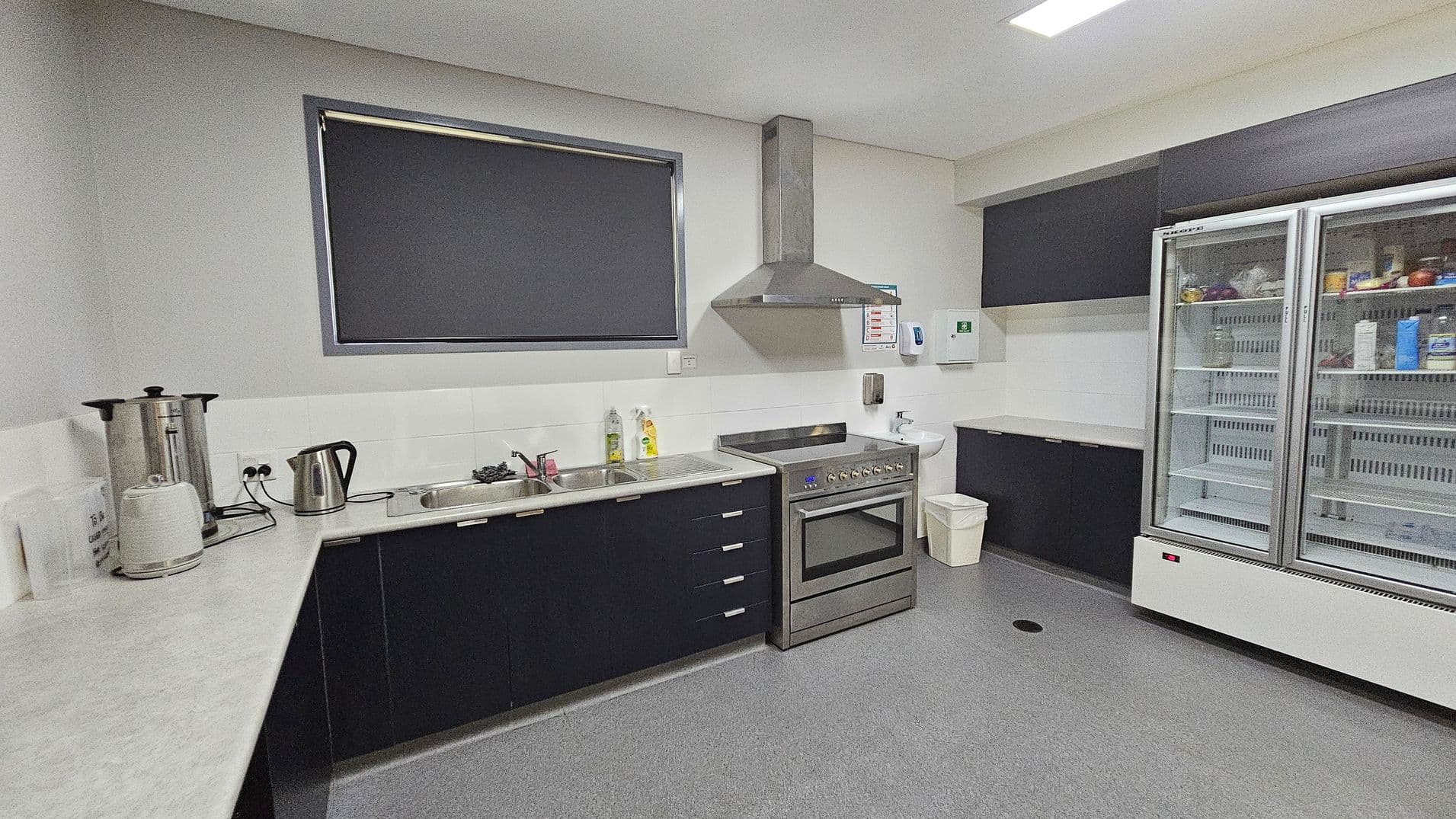 A modern kitchen with grey flooring and white walls. On the left, there is a long countertop with a large kettle, a smaller electric kettle, and a toaster. A window with a dark blind is above the sink. In the centre, there is a stainless steel oven with an extractor hood. To the right, a large double-door fridge with glass doors contains various food items. Above the fridge, there are dark cabinets. A small sink and a bin are near the oven.