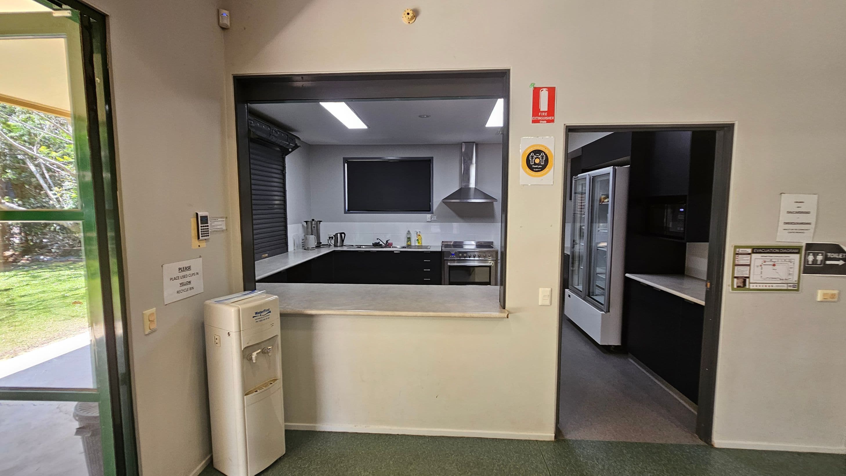 A kitchen area with a serving window in the centre, featuring a countertop, stove, and range hood. To the right, there's a doorway leading to a room with a large refrigerator. On the left, a water cooler is positioned near a partially open door showing greenery outside. The floor is green, and there are various signs on the walls, including a fire extinguisher sign. The ceiling has recessed lighting.