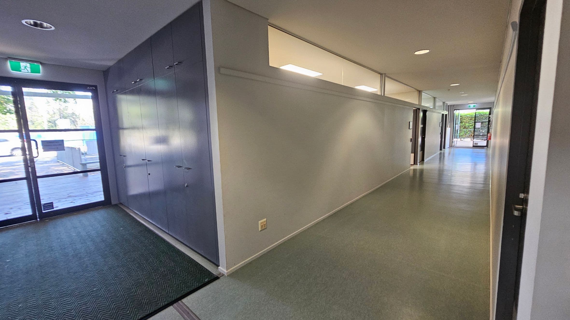 A hallway with a grey and green floor. On the left, there is a glass door with an exit sign above it and a row of grey lockers. The right side features a long white wall with windows near the ceiling, and several doors leading to rooms. The ceiling is white with recessed lighting. At the far end, another glass door is visible, leading outside.