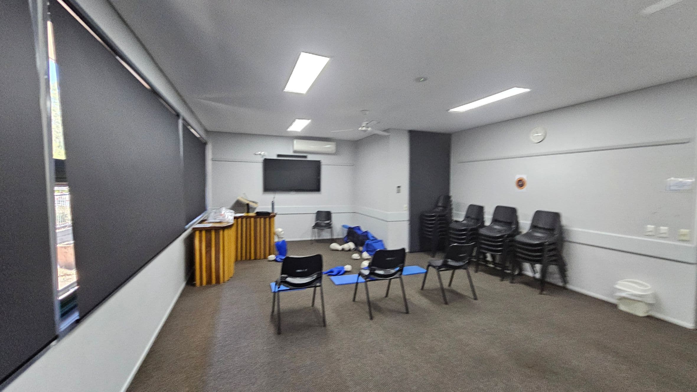 A room with grey walls and a carpeted floor in shades of green and brown. On the left, there are large windows with black blinds. In the centre, several black chairs are arranged facing a wall-mounted TV screen. Blue mats and CPR mannequins are scattered on the floor. On the right, stacks of black chairs are piled against the wall. A wooden lectern is positioned near the windows. The ceiling has recessed lighting and a ceiling fan.