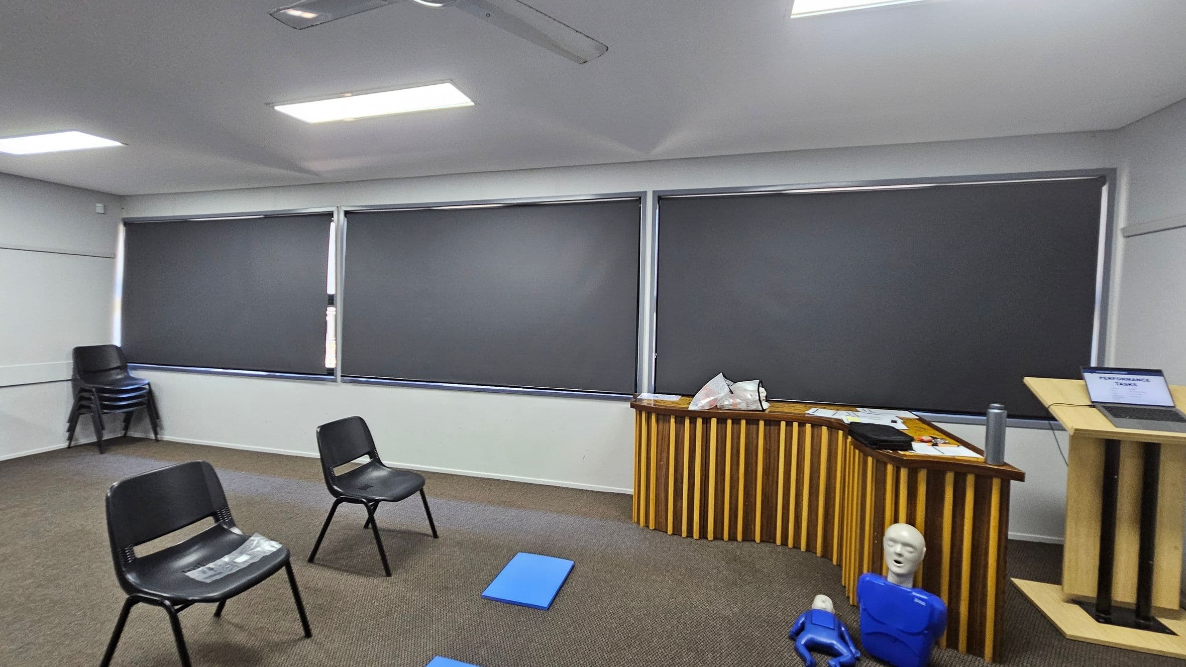 A room with grey walls and a ceiling with fluorescent lights. On the left, there are stacked black chairs against the wall. In the centre, two black chairs are spaced apart on a carpeted floor with green and brown patches. On the right, a wooden podium holds a laptop displaying "PERFORMANCE TASKS." Nearby, a wooden table has papers and bags on it, with two blue mannequins on the floor in front. The windows are covered with dark blinds.