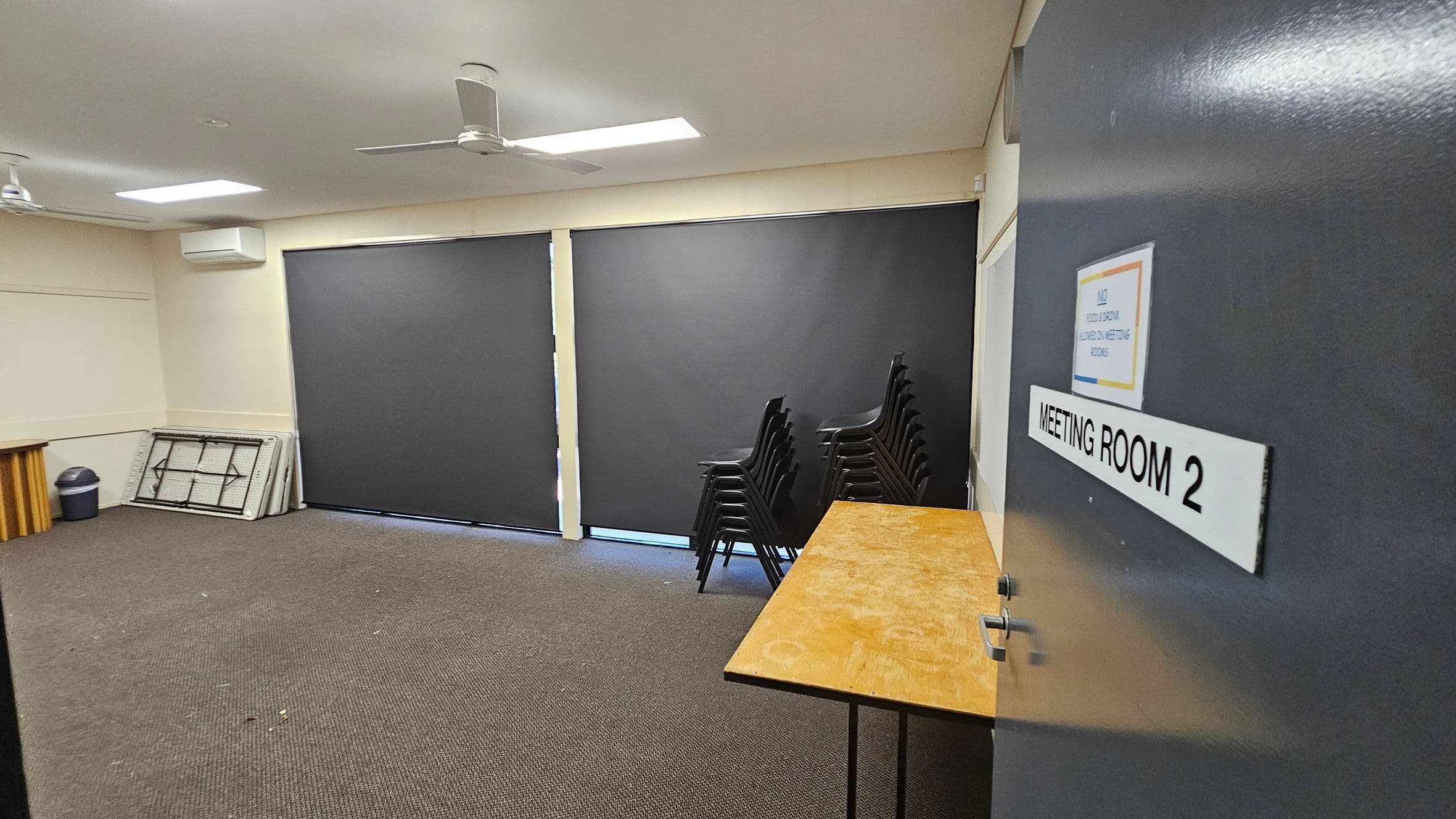 A meeting room with a brown carpeted floor and a white ceiling featuring two ceiling fans. On the right, a door labelled "MEETING ROOM 2" is partially open. Inside, a wooden table is visible. Against the far wall, there are large black blinds covering the windows. To the left, a stack of folded tables and a bin are placed in the corner. A stack of black chairs is positioned near the centre of the room.