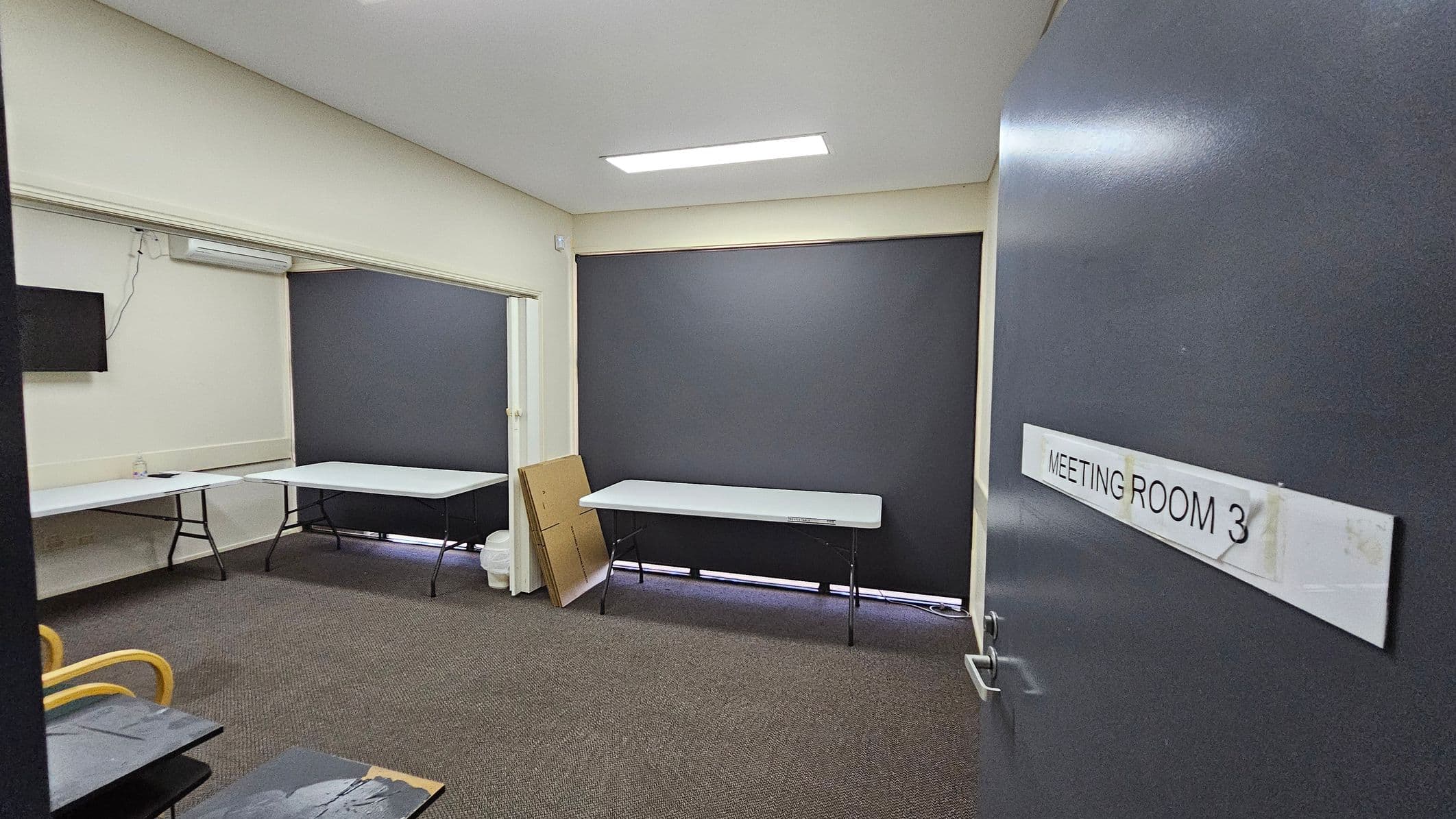 A room with grey walls and a carpeted floor. On the right, a door is slightly open with a sign reading "MEETING ROOM 3". Inside, there are two white folding tables against the walls. A small television is mounted on the left wall. A stack of cardboard and a bin are near the back wall. The ceiling has a rectangular light fixture. The windows are covered with dark blinds.