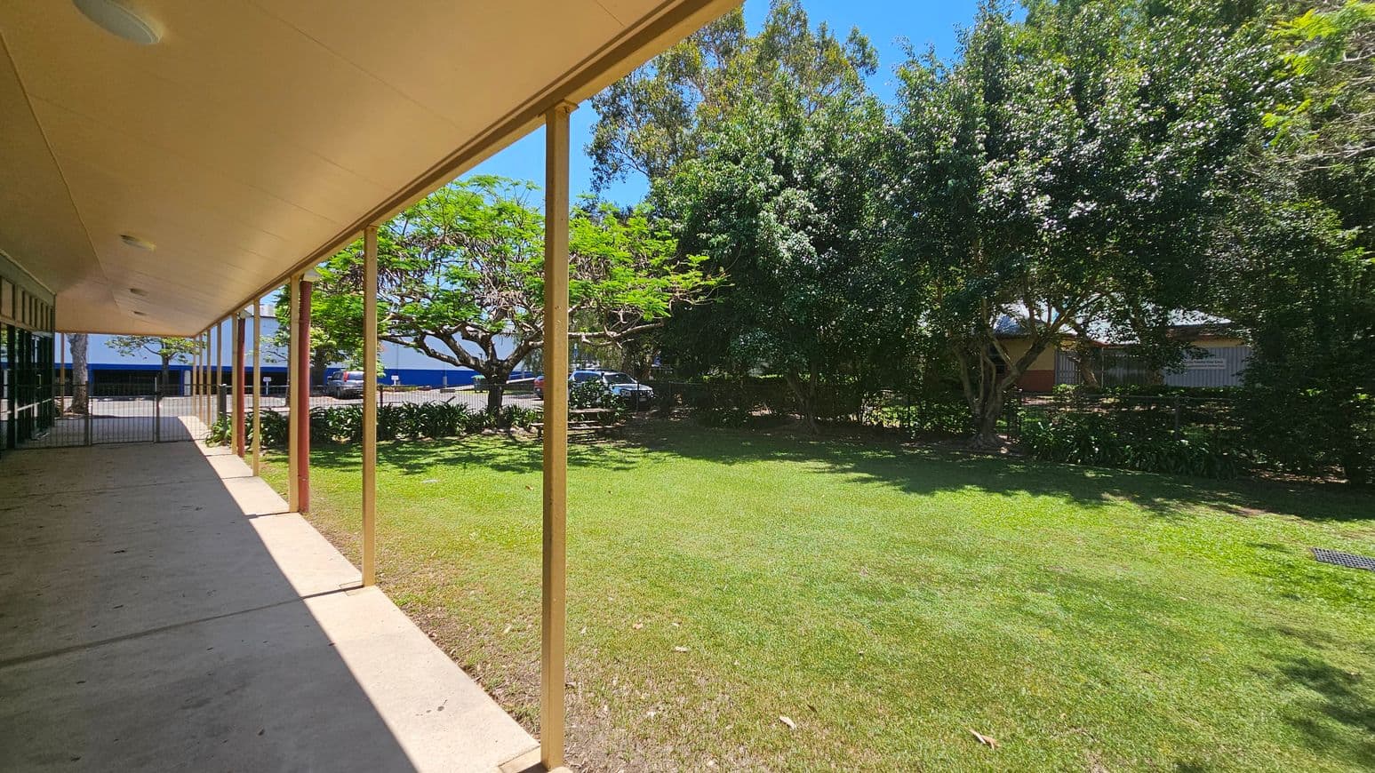 A covered walkway with a green wooden wall on the left and glass doors. A fire hose reel is mounted on the wall. The concrete path extends forward, bordered by a grassy area with a large tree on the right. Several parked cars are visible in the background. The ceiling is beige with round lights, and wooden posts support the roof.