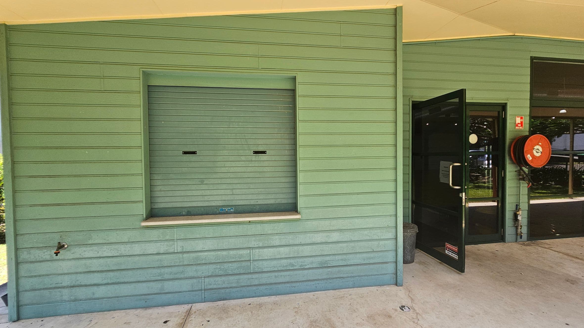 A green wooden wall with horizontal panels. On the left, there's a closed service window with a small ledge and a tap below. On the right, a black-framed glass door is open, leading inside. Next to the door, a red fire hose reel is mounted on the wall. The floor is concrete, and the ceiling is light-coloured.