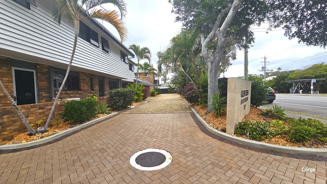 To the right is a road. A landscape of plants and concrete signage bearing the facility name stands before the entrance. To the left is a two-storey building with a facade combining brown bricks and corrugated design. In the middle is a driveway with bricks. A drainage hole with a cover is visible in the middle. The left and right are bordered by a landscape of plants, orange pebbles, palm trees, and grass.