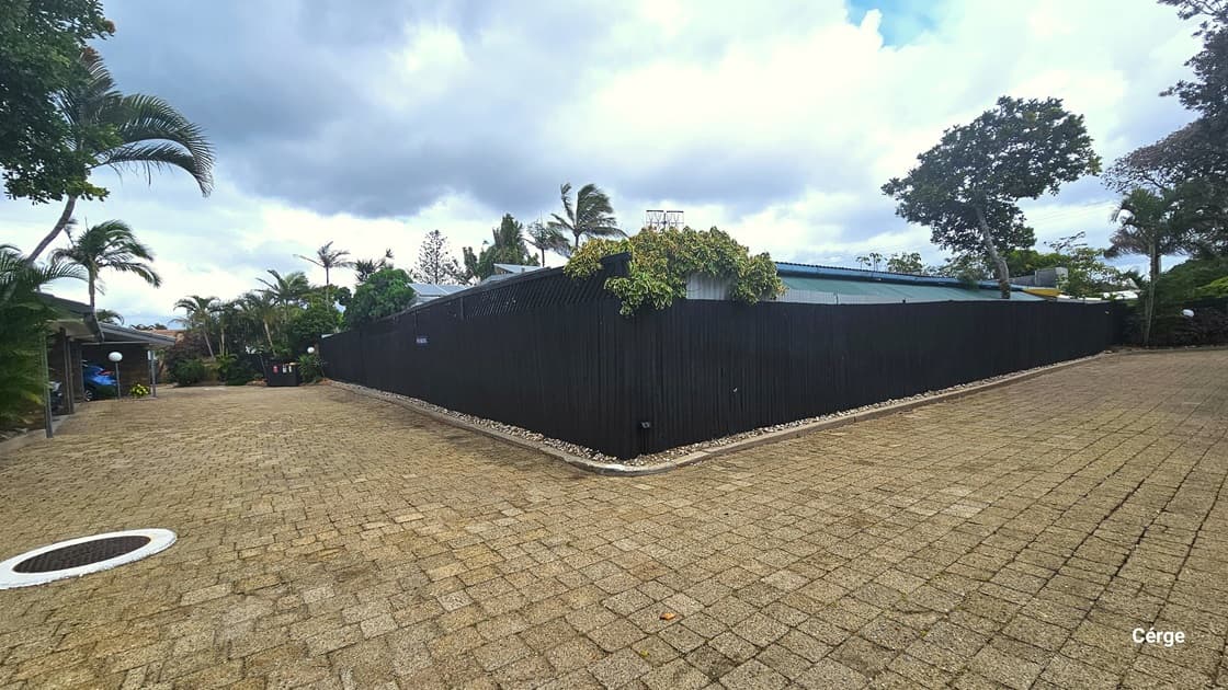 The driveway curves at the corner. A black metal fence borders the road. Brick designs adorn the driveway. A landscape of plants and palm trees line the road on the left. A drainage hole with a cover sits on one side of the driveway on the left.