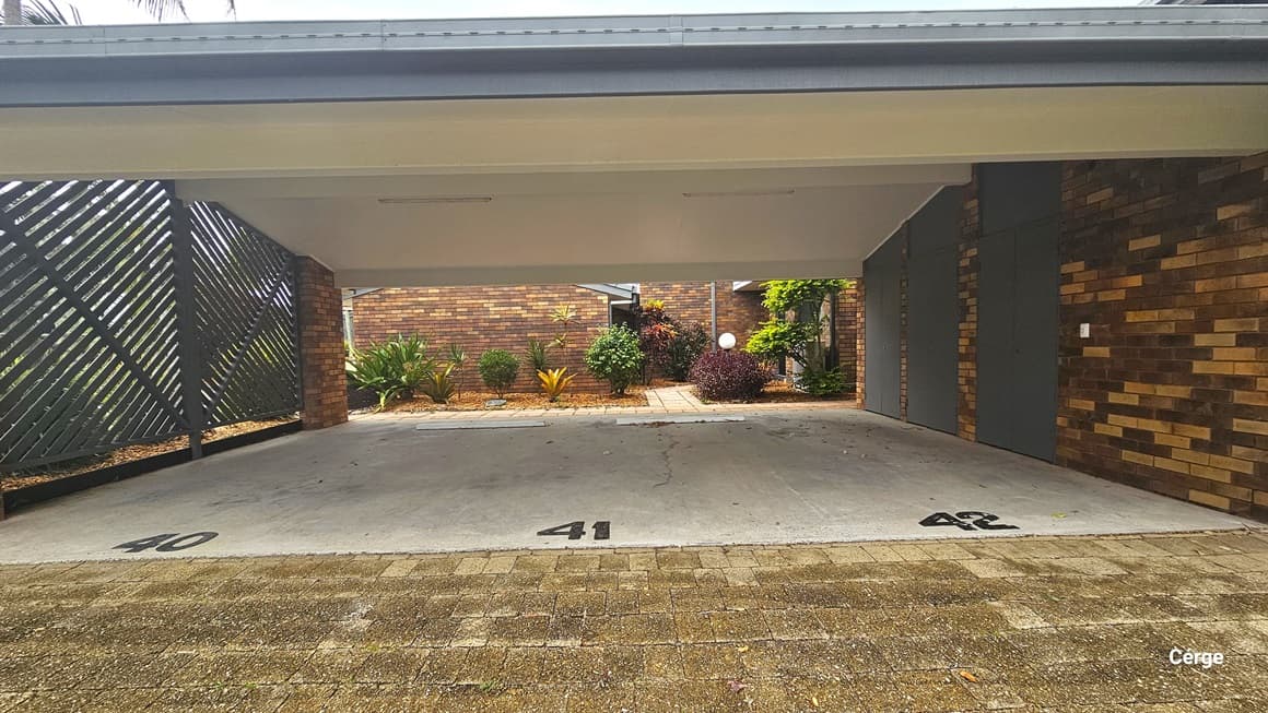 The low-ceiling undercover parking has markings indicating the parking slot numbers. A grey metal fence secures the left, while on the right is a brick wall. Behind the parking is a building with a brick facade. Landscape gardens decorate the front. The floor transitions from bricks to concrete.