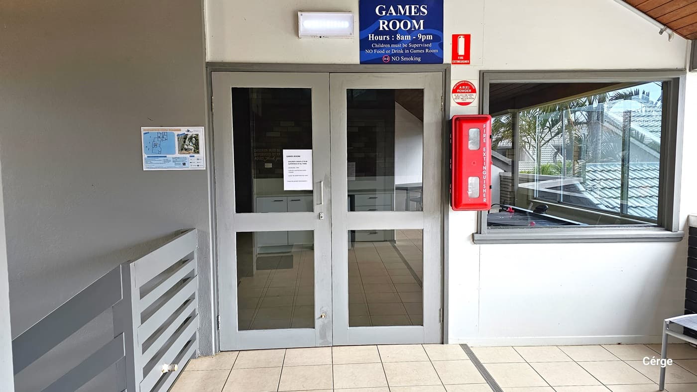 A manual outward-opening glass door. Reminder posters and fire extinguishers are mounted on the wall. To the left is a stair with handrails and a seating area to the right. The floor is tiled in white, the walls combine white and grey, and the ceiling is dark timber.