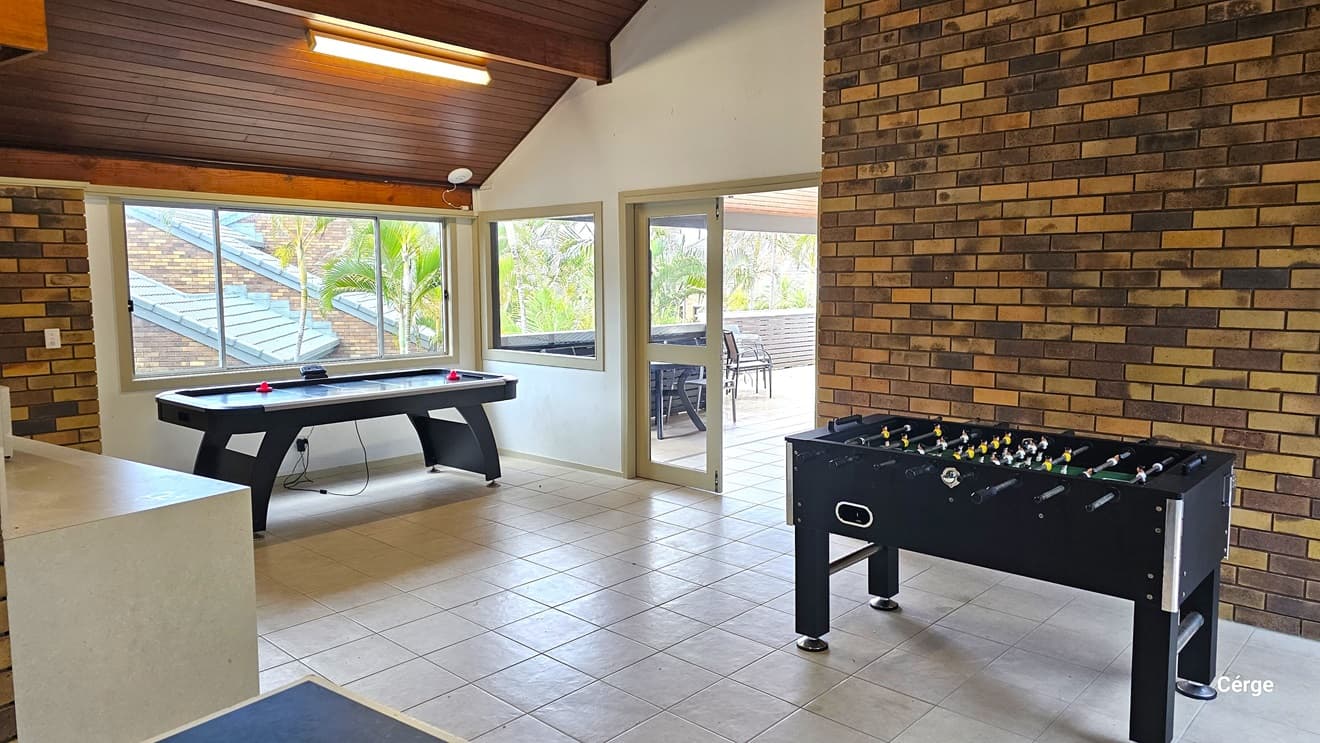 To the right, a foosball table and a table near the wall. On the left, an air hockey table is positioned by the glass windows. Adjacent, is a small kitchen area. The ceiling features brown timber and fluorescent lights, with brown brick walls. The floor is tiled in white. Outside the manual glass door, tables and chairs are arranged on the balcony overlooking palm trees and buildings.