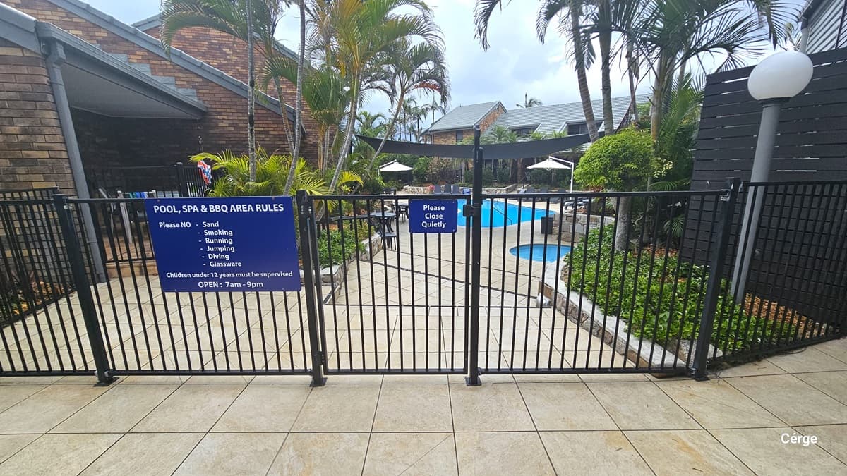 On the right, a concrete building with a grey wall, featuring a wooden chair against it. Next to it, a wooden wall and a metal gate with blue signages and reminders. To the left, is a landscape of plants and palm trees in planter boxes and a lamp post. Beyond the gate lies a building with a brick facade and a pool.
