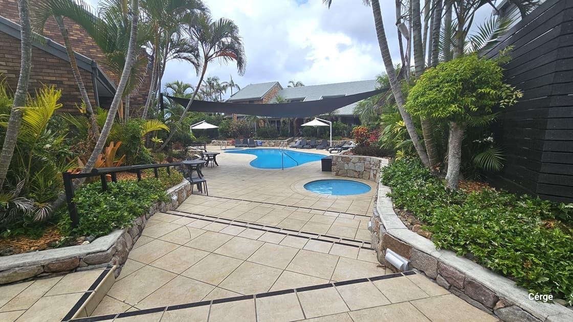 Steps lead to the pool area furnished with tables, chairs, and shaded spots. On the right, is a small circular pool, with the main pool beyond, offering steps with handrails and a ladder. To the left, is a building with a brown brick facade. Landscaped garden with plants, palm trees, and wooden railing on both sides. A black shade cloth hangs above. Another building stands behind the pool.