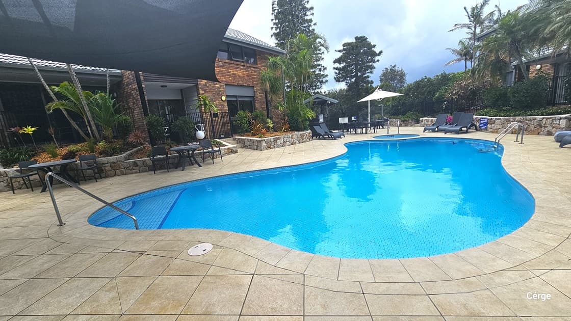 In the middle lies a large pool, complete with steps, handrails, and a ladder, with depths from 1.1m to 1.8m. To the left stands a building with a brick facade. Surrounding the pool area is a landscaped garden of plants and palm trees. Tables, chairs, and sun loungers are arranged on the pool deck under shades. Overhead, a black shade cloth provides cover for a section of the pool. To the right, another building is bordered by trees and plants.