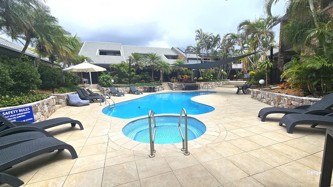 In the middle is a spa pool and the main pool, both with steps and handrails. Tables, chairs, and sun loungers line the pool deck. Shade cloths and umbrella shades provide cover. Buildings with brick facades and corrugated metal roofing, along with landscaped gardens and palm trees, surround the pool deck. Blue posters displaying safety reminders are also visible.