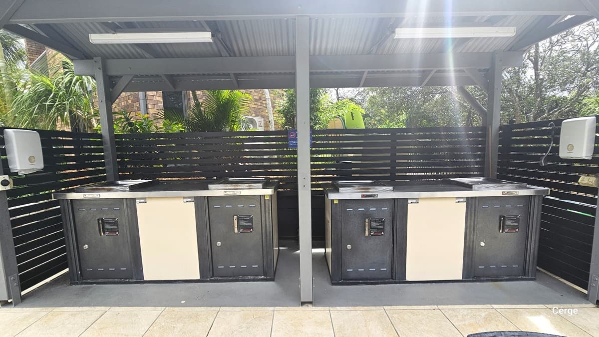 An open area shaded by corrugated roofing, featuring four barbecue grills underneath. A metal fence surrounds the space, with tissue dispensers mounted on the left and right walls. Fluorescent lights hang from the ceiling. Behind is a building with a brick facade, bordered by palm trees and tall plants.