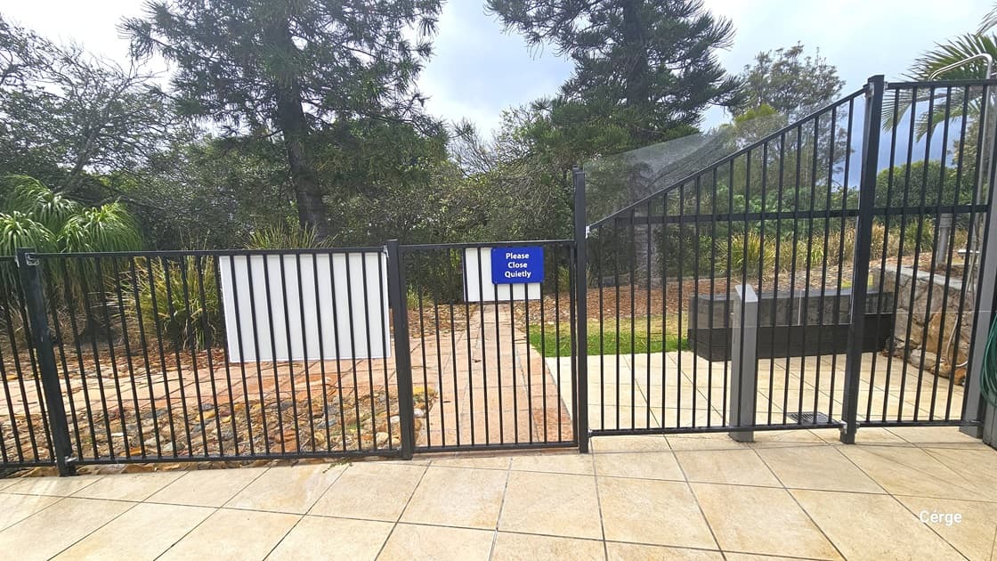 A black metal fence and gate are equipped with blue signage bearing reminders. Behind the gate, there is a concrete pathway leading to the garden and beach area. Tall trees, rocks, and grass border the path.
