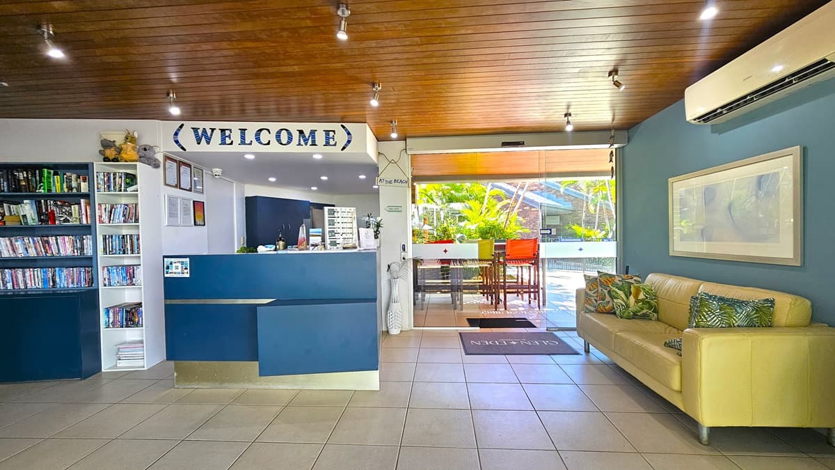 On the right side, a reception desk with a blue front displaying a "Welcome" sign. Framed posters and certificates adorn the wall nearby. Adjacent, a display rack holds games, books, and DVDs for hire. A yellow couch rests against the green wall, topped with a framed decoration and an air conditioning unit. A glass sliding door leads to additional seating outside. The ceiling boasts dark brown timber with light fixtures, while the floor is tiled in white.