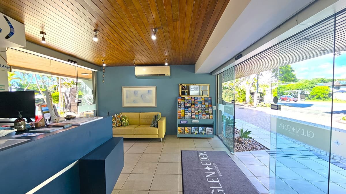 To the left, a reception desk with a blue front, topped with a computer and brochures. To its left, is a glass sliding door. A yellow couch against the blue wall, adorned with a poster and air conditioning. Adjacent, a display rack offers books and DVDs for rent. Dark brown timber lines the ceiling with light fixtures. The floor is tiled in white, with a grey mat at the doorstep. An automatic glass sliding door is on the right. Outside, the driveway and road are lined with trees and plants.