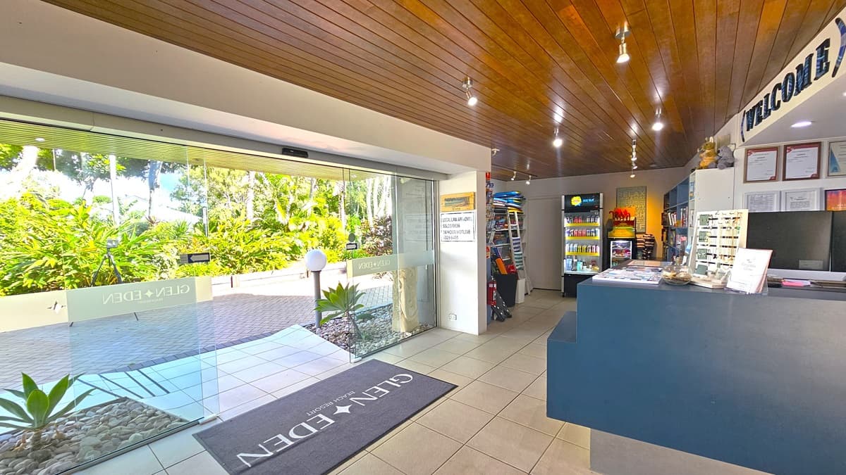 To the right, a reception desk with a blue front, topped with a computer and brochures. To its right, is a fridge displaying cold drinks and a display rack filled with toys and accessories, along with a fire extinguisher Dark brown timber lines the ceiling with light fixtures. The floor is tiled in white, with a grey mat at the doorstep. An automatic glass sliding door is on the left. Outside, the driveway and road are lined with trees and plants.