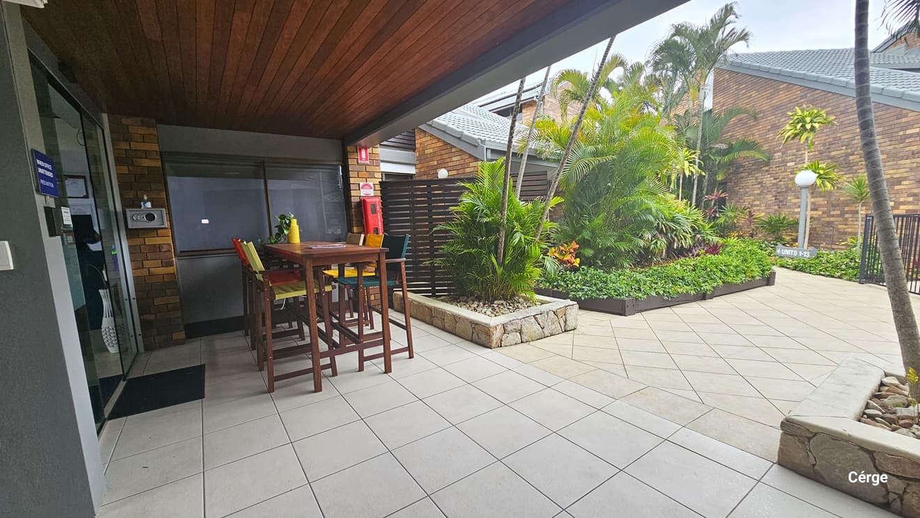 On the left, is a glass sliding door with a grey mat. A wooden table and high chairs are arranged nearby. A fire extinguisher is mounted on the brick wall. The ceiling is adorned with brown timber, and the floor is tiled in white. To the right, a garden with landscaped plants and palm trees in planter boxes. A building with a brick facade stands on the right. A garden lamp and wooden fence are also visible.
