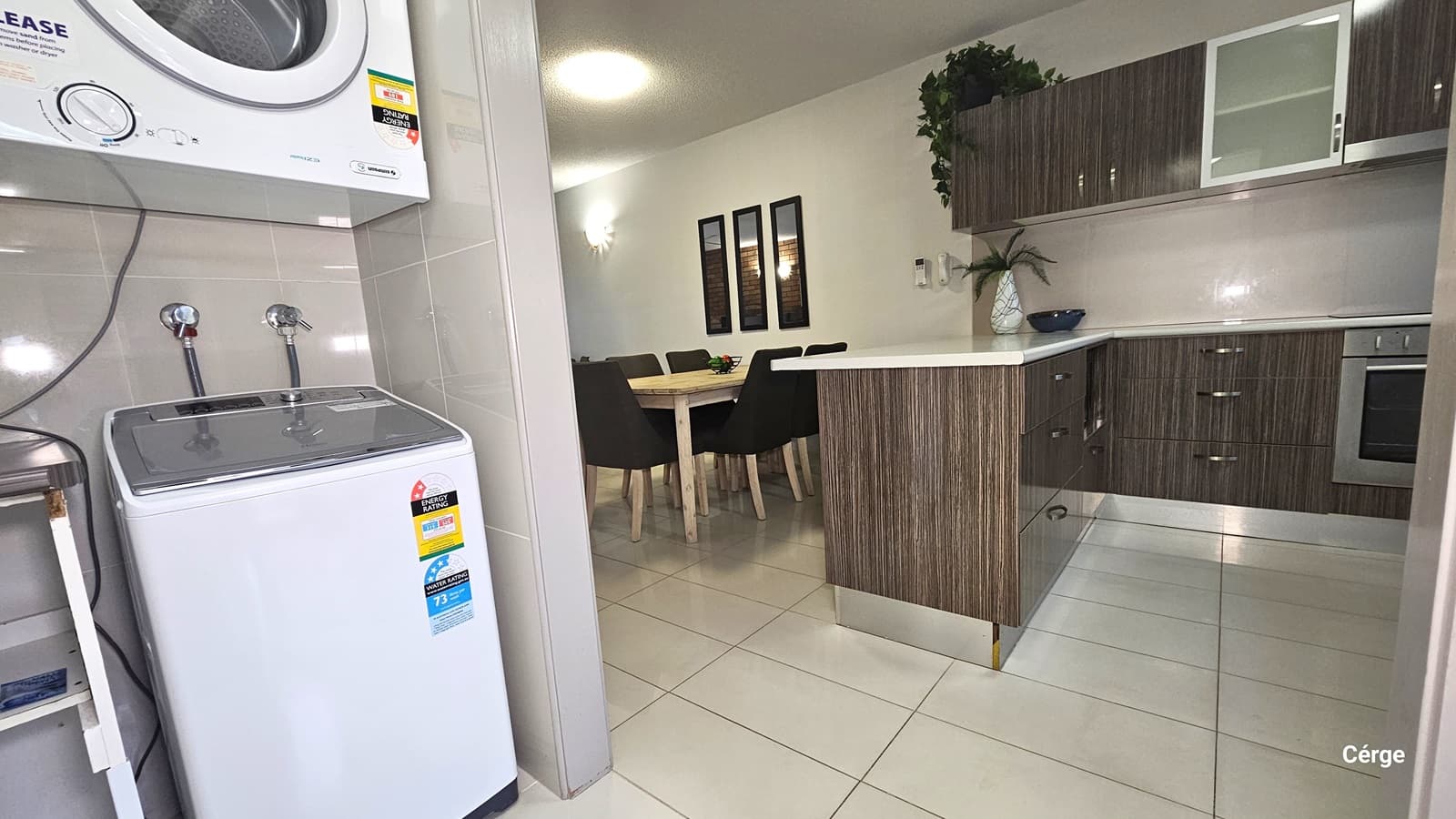 To the left, a washing machine sits, with a clothes dryer mounted overhead and a table beside. On the right, a kitchen area is equipped with cabinets designed with brown timber, holding a flower vase and foliage decorations. Adjacent is a dining set. Three vertical mirrors are hung on the wall, with light fixtures on the right. The ceiling is white, and the floor is tiled in white.