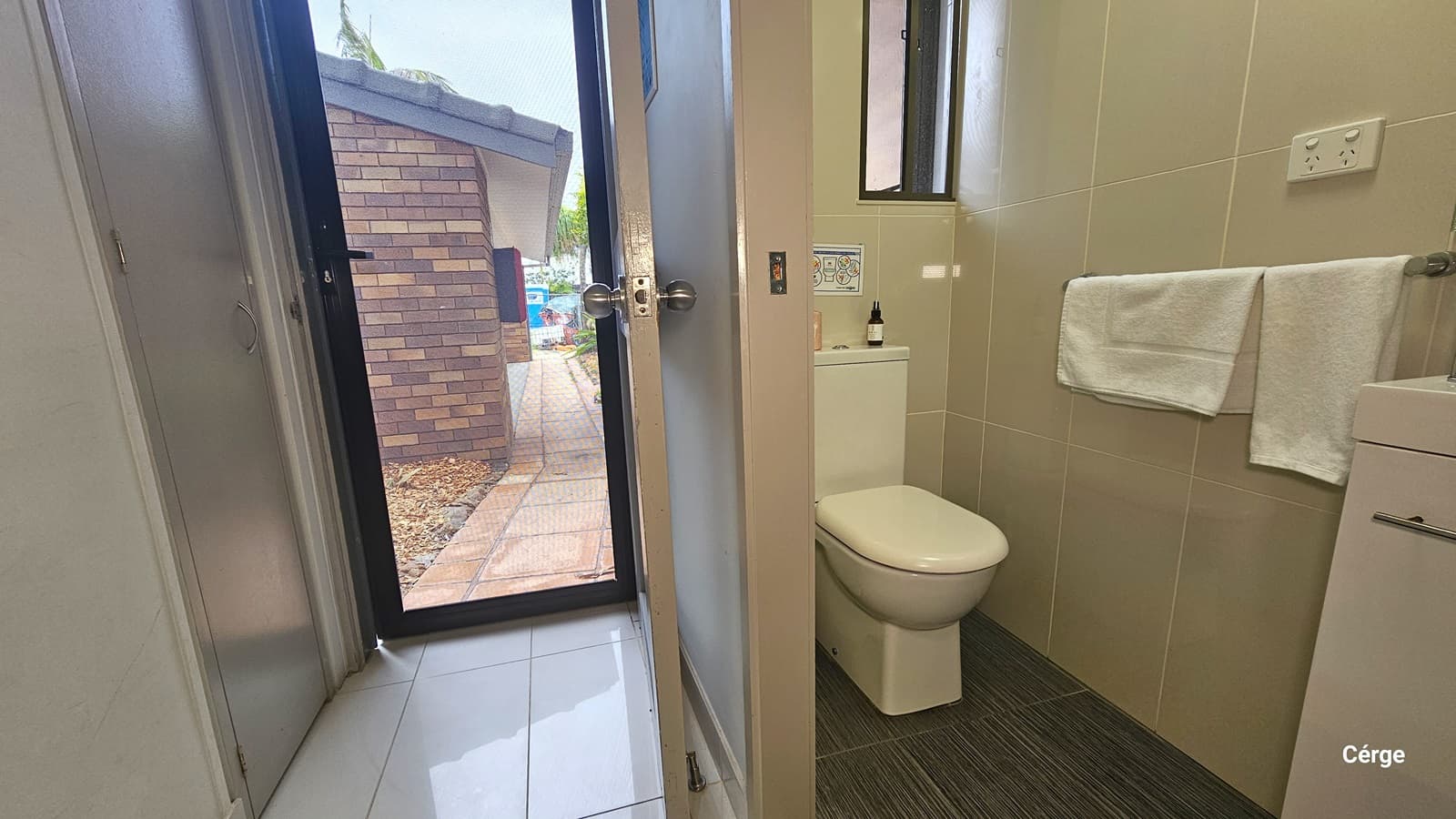 Inside the small toilet. A toilet bowl is positioned in the corner. Adjacent is a handrail equipped with white towels and a sink with a cabinet. The wall features cream tiles, and the floor has dark grey tiles. To the left is a door serving as an entrance, equipped with a screen door offering a view of the brick building and pathway outside. The floor has white tiles.