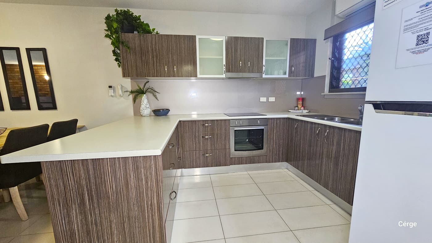 The kitchen comprises a kitchen island with a brown timber design, cabinets, and a cooktop. A sink is positioned to the right, with a window above fitted with wire mesh. A fridge stands to the right. A dining set is positioned to the left. The floor is tiled in white, and the ceiling and walls are white.