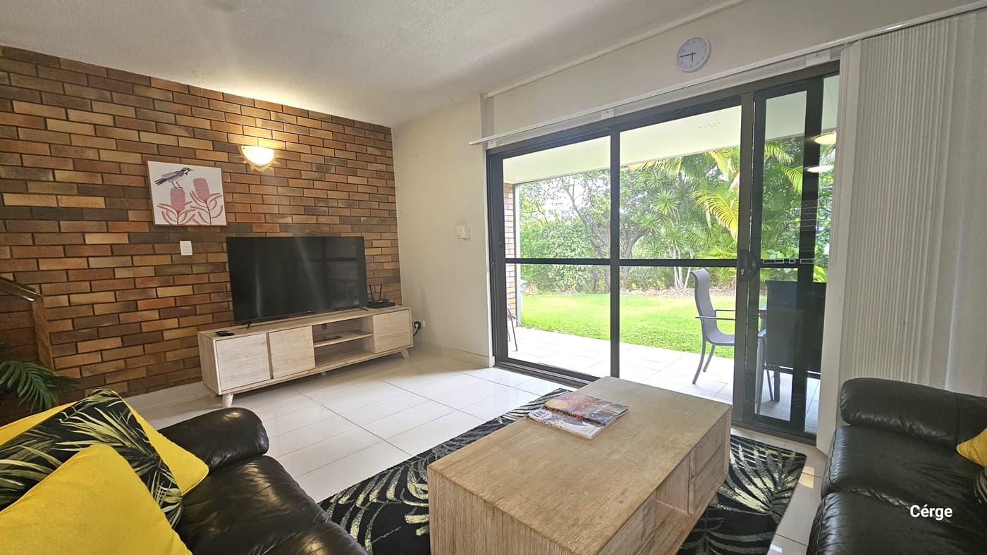 The living area comprises black leather couches with colourful pillows. A wooden centrepiece holds a magazine on top of a carpet. To the left, a flatscreen TV sits on a TV stand with a timber design. A painting and a light fixture are mounted on the brick wall. To the right, a glass door with white curtains drawn to the sides. A round clock is mounted on the upper middle wall. Outside, greenery is visible through the glass door.