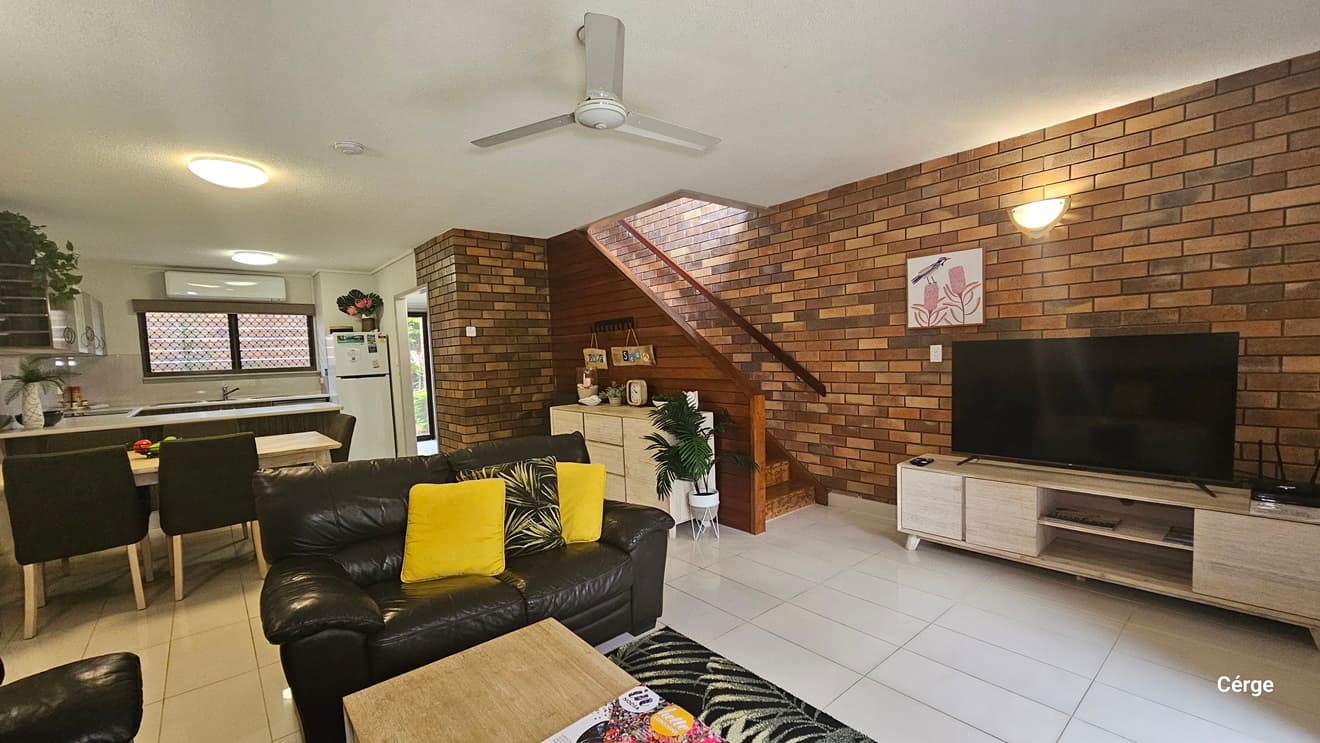 The living area features black leather couches with colourful pillows. A wooden centrepiece holds a magazine atop a carpet. To the right, a flatscreen TV is on a TV stand with a timber design. A painting and light fixture adorn the brick wall. On the left, is the kitchen and dining area. To the right, is a display cabinet, and behind it is a stair with handrails. White tiles cover the floor, while the ceiling is white with a ceiling fan and LED lights.