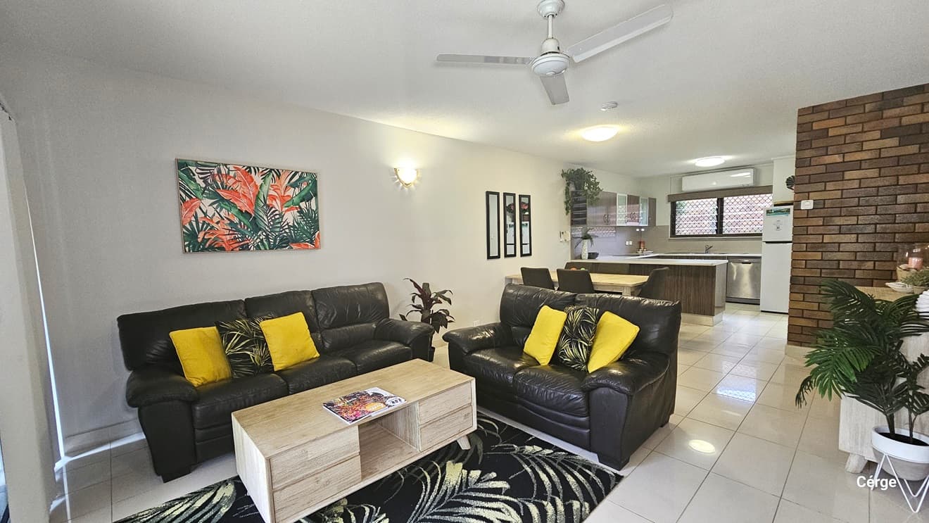 The living area has black leather couches with colourful pillows. A wooden centrepiece holds a magazine on a carpet. Adjacent, a dining table and the kitchen area with foliage decoration hanging from the cabinet and three mirrors mounted on the wall. To the right, a display cabinet holds decorations. White tiles cover the floor, and the ceiling is white with a ceiling fan and LED lights.