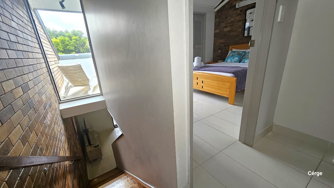 To the left, there is a stair with handrails mounted on the brick wall. On the right, there is a white wall partition. Behind the partition is a door that leads to a bedroom with a queen-size bed. Colourful pillows and rolled white towels sit on top of the bed. The floor has white tiles, and the wall inside the room is adorned with brown bricks.