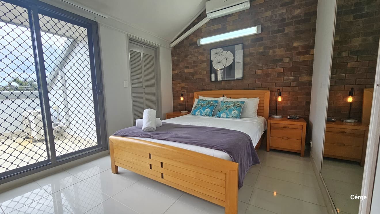 A queen-size bed with a wooden headrest. Bedside tables with light fixtures on both sides. A floral painting, a fluorescent light, and an air conditioning unit mounted on the brick wall. To the right, a large mirror; to the left, a floor-to-ceiling glass door with a metal mesh screen leading to the balcony. A wardrobe stands beside it.