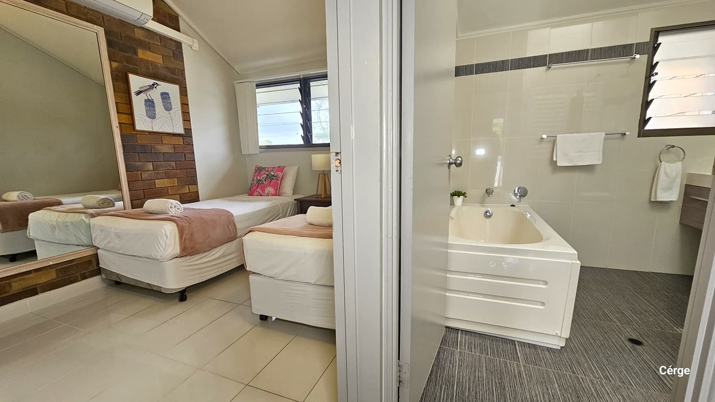 To the left is a room with two single beds. A bedside table holding a lampshade sits between the beds. A large mirror and wall decoration are mounted on the brick wall. A window overlooks the beds. To the right is a bathroom. A tub is positioned against the left wall, with hand grabs and white towels mounted on the wall. A washbasin is on the right near the window. The floor changes from white tiles in the bedroom to grey tiles in the bathroom. The ceiling and walls are white.