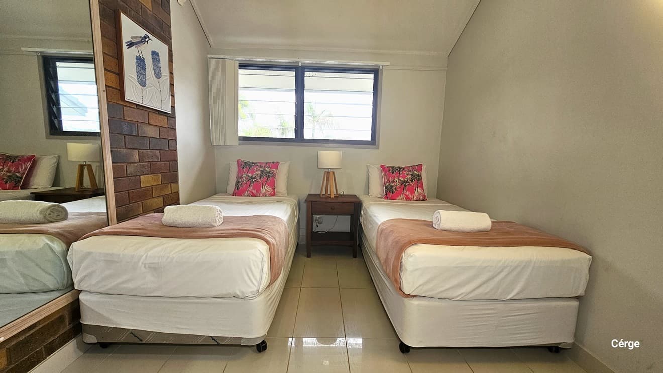 Two single beds with white and brown mattresses and white and bright pink pillows. A bedside table is placed between them with a lampshade on top. Above the beds, a window with a white curtain pulled to the left. A large mirror and wall decoration are mounted on the brick wall to the left. The floor has white tiles, and the ceiling and the right wall are white.