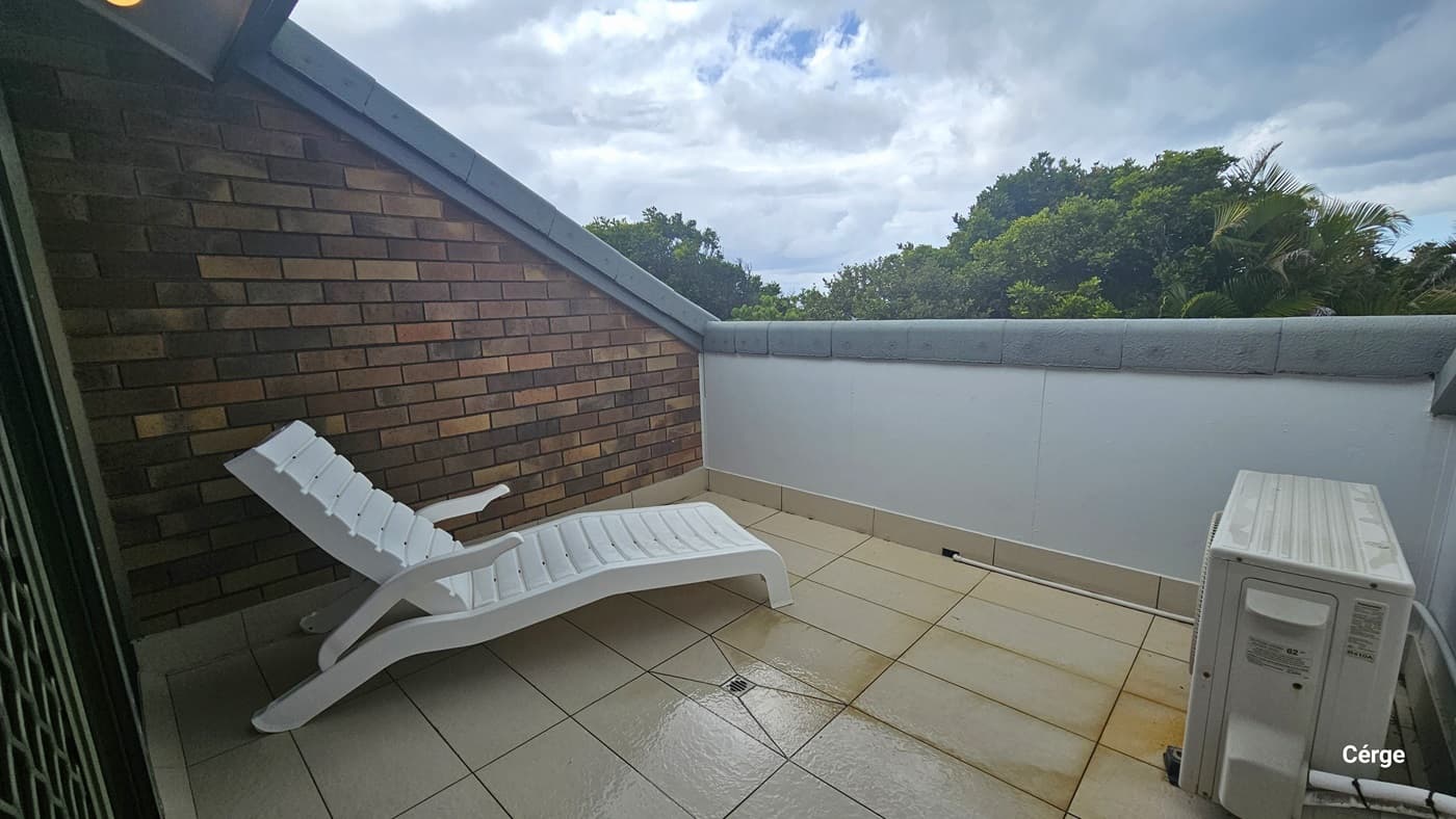 The balcony features a white sun lounger chair on the left near the brick wall, with a condenser unit on the right. Secured by a concrete wall, trees are visible from outside. The floor has white tiles with rust stain markings.