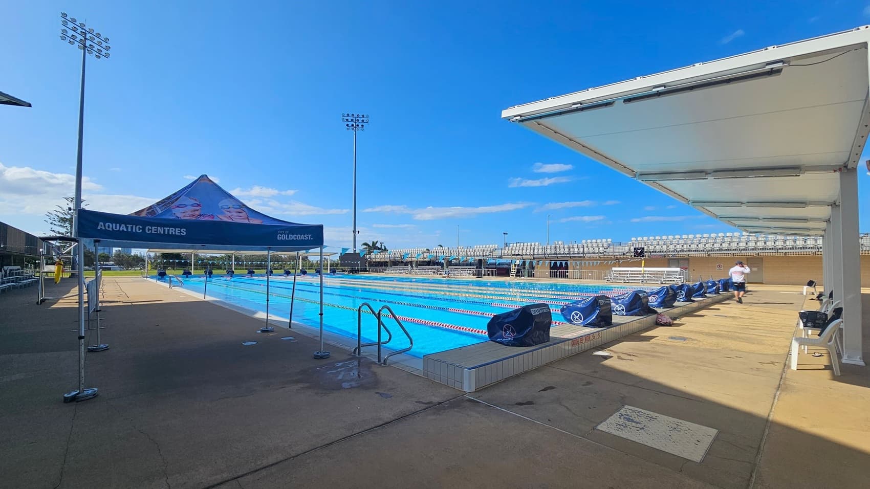 An outdoor swimming pool with multiple lanes, surrounded by a concrete deck. On the left, a white plastic chair is placed near the pool's edge. In the background, there are tall buildings and a few trees. The pool is covered by a partial canopy, and there are stadium-style seating areas on both sides. Floodlights are mounted on tall poles, and a digital clock is visible on the right side. The sky is partly cloudy.